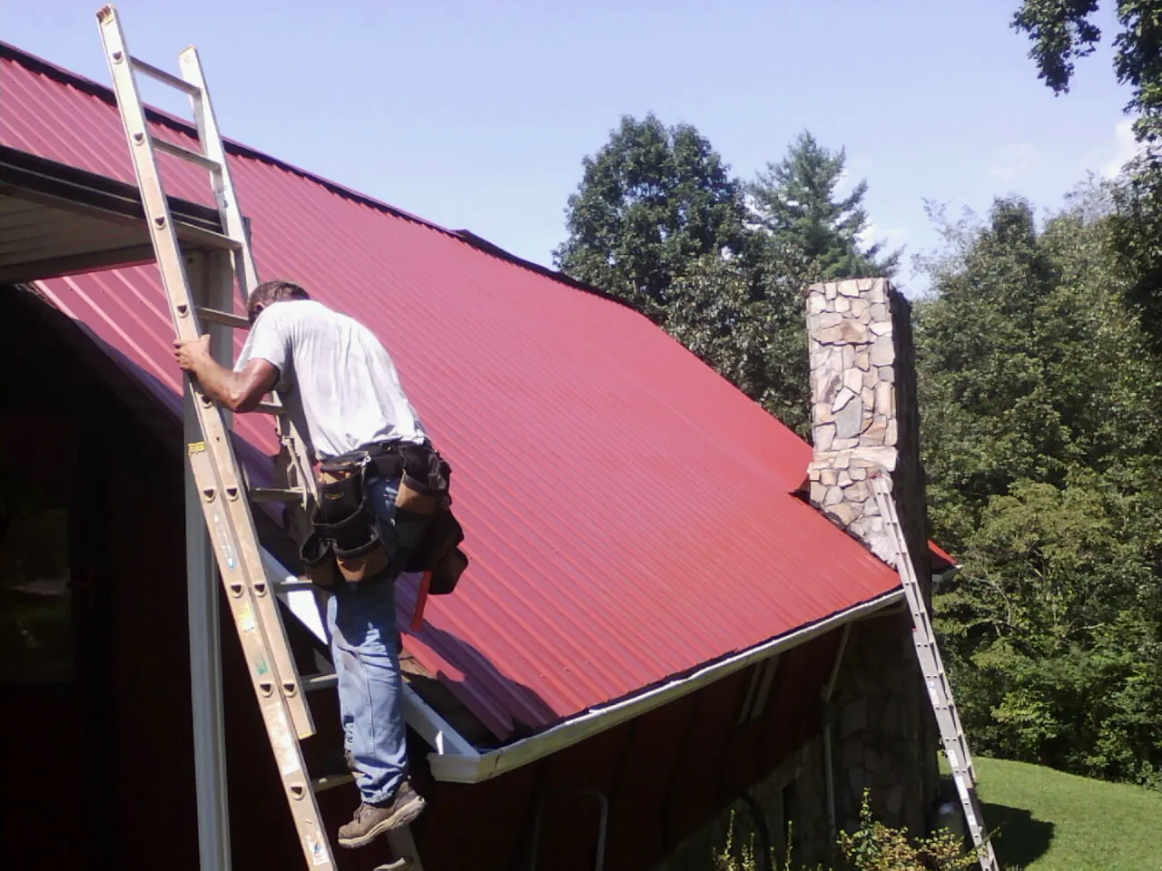Roofer on a ladder beside a red metal roof and stone chimney — Soffit & Fascia Repair by Roofing Brothers in Longbranch
